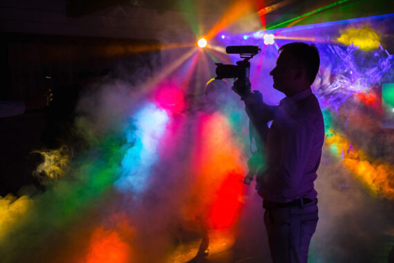 A videographer is on stage amongst multicoloured lights and fog from a dry ice fog machine.